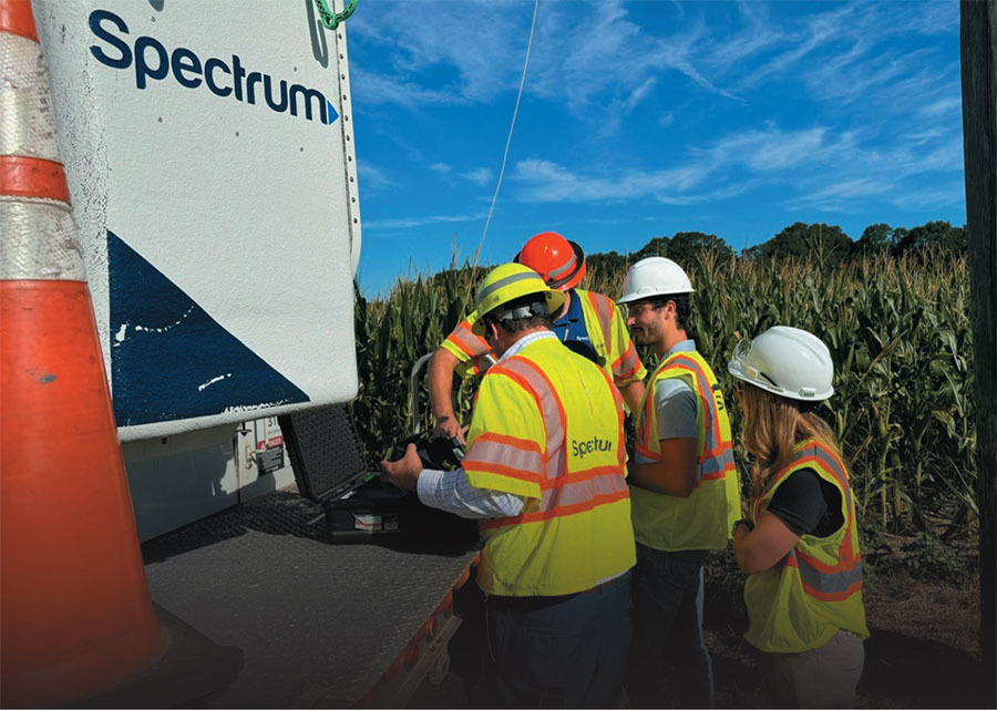 A group of Spectrum workers in safety vests and hard hats review equipment beside a service truck near a cornfield.