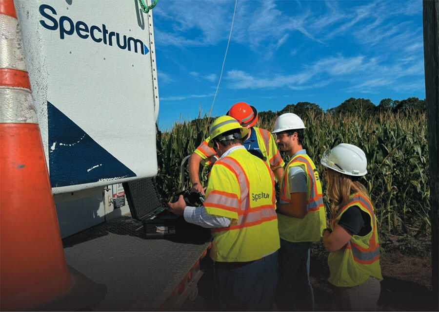 A group of Spectrum workers in safety vests and hard hats review equipment beside a service truck near a cornfield.